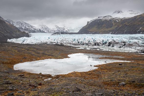 Landscape photo of the Svinafell glacier. Foreground frozen puddle of meltwater, surrounded by soft moraine. Background of snow covered mountains