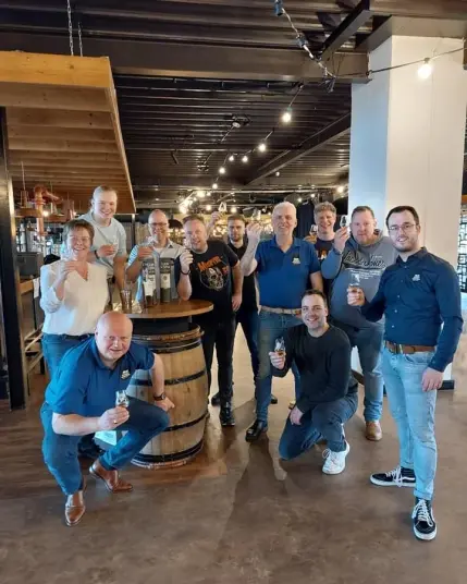 De mensen van de organisatie van het Whisky Weekend Twente met een aantal vrijwilligers rondom een vat waarop de festivalbotteling staat. De organisatie heeft donkerblauwe poloshirts aan met het logo van Whisky Weekend Twente.