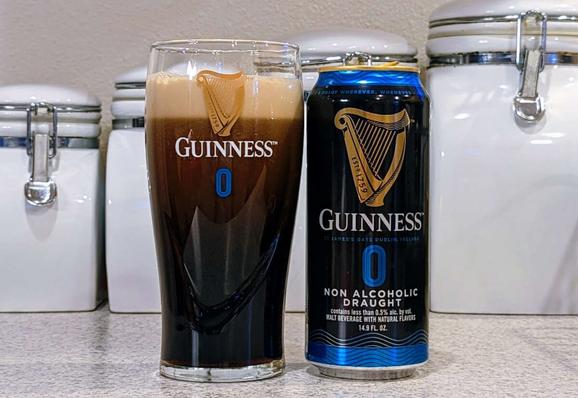Tall glass of Guinness 0, the non-alcoholic version of Guinness Stout draught on nitro. It's a dark stout brew with a creamy tan head of foam, in a branded "Guinness 0" glass, next to the can it was poured from.