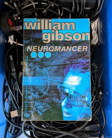 Image shows a blue book, the cover featuring hazy pictures of technology and a man's face wearing futuristic glasses/goggles. The words "William Gibson" and "Neuromancer" are at the top. The book rests on a tangled bunch of black cables inside a blue box.