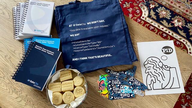 A table full of notepads, tote bags, cookies, stickers from DataCite. On the right a sheet of paper with an abstract picture of a greek thinker thinking about PID (Persistent Identifier).
