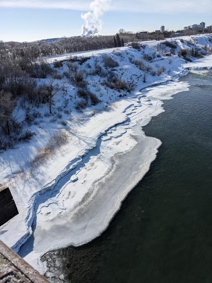 Photo of a river bank with ice and snow. Taken from a bridge looking towards the bank