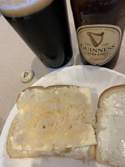 A paper plate with a sandwich on it consisting of white bread, mayo, spicy brown mustard, thick slices of Limburger cheese, and sauerkraut. In the background are a bottle of Guinness Extra Stout, the bottle cap, and a glass containing the bottle’s contents.
