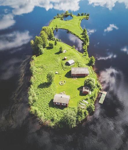 Same as island but in summer. Looks like it is floating in the sky (which is a near mirror reflection of deep blue puffy clouds).  The vegetation is all a bright and vibrant green. There are three, nearly hidden canoes laying keel up in the lower right. Slightly above that is a brand new L-shaped floating dock with a green fishing net spread across it, drying in the sun.