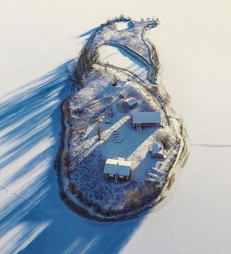 The island in winter. The surrounding water and the pond / inlet are iced over. There is a light coat of snow on everything. There are long shadows slanting down on the left across the ice from the sun shining through the leafless trees and shrubs.