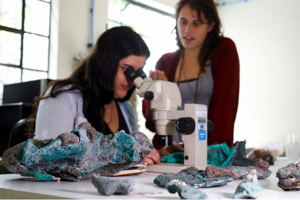 Female researcher in a lab coat looks through a white microscope at blue-ish rocks as another woman in red looks on.  More "rocks" scattered about the table.  many are blue or appear to be normal rocks glued together by blue.  The blue is melted plastic that has cooled.