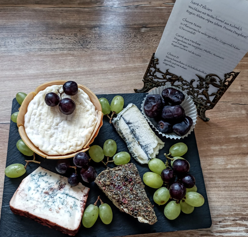Auf einer schwarzen Schieferplatte die 4 Sorten Käse, Weintrauben und Datteln. Daneben steht ein kleiner antiker Fotoständer, darin ein Blatt Papier, auf dem die Namen der Käse und ihre Herkunft aufgeschrieben sind
