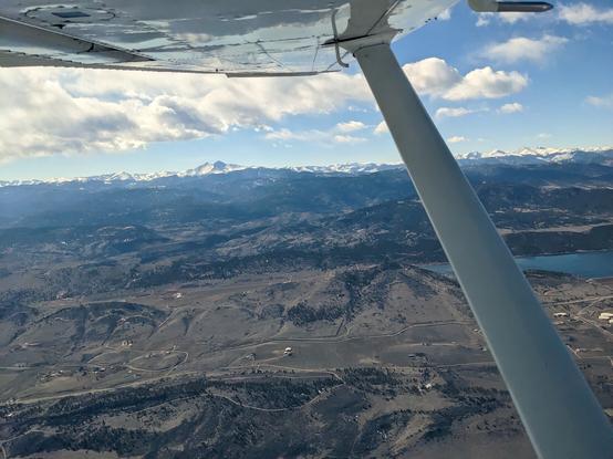 View along the underside of the left wing of a Cessna 182 with the wing strut running from the bottom right corner of the frame diagonally to the wing which overs the top 1/5 of the frame.

The ground below shows the foothills of Colorado with Horsethooth reservoir in the midground. In the far distance is snow covered Longs Peak with a swath of blue sky above and then clouds.