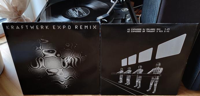 Eine Schallplatte wird auf einem schwarzen Dual-Plattenspieler abgespielt, davor steht die schwarze Plattenhülle mit dem weißen Computerschruftzug "KRAFTWERK EXPO REMIX" und dem weißen "Impuls"-Fraktal-Logo der #Expo2000 auf dem Cover. Rechts daneben ein schwarzes Plattencouvert mit einer weißen Grafik in schräger Perspektive der Musikgruppe Kraftwerk, die in Drahtgitteranzügen unter ihren 4 Synthesizern stehen, hinter ihnen an der Wand 4 graue 16:9-Bildschirme
