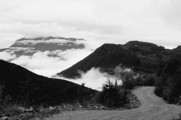 Unpaved, winding gravel backroad.  Big clouds & mountains in the background. Black and white photo.