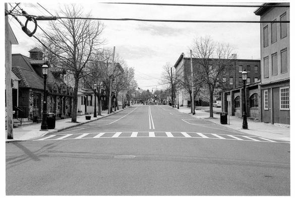 A black and white photograph of a downtown business district during the daytime, taken from the middle of the road with no signs of human activity. Beyond the first intersection in the distance, the center of town with a public clock and some trees, and behind that, an overpass for elevated commuter rail tracks. An American flag is visible over tracks, from the flagpole by town hall.