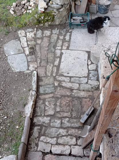 Vue plongeante sur une allée pavée avec de la pierre d'écurie. Le chantier est presque terminé.
En bas à droite, appuyés contre un escalier de pierre, un balai, une pelle et une planche. Plus haut, un chat noir et blanc, un seau de graviers, une cagette d'outils. En haut de l'allée pavée, devant la première marche, trois grosses dalles. Le pavage s'ouvre en éventail pour amorcer le chemin qui part de la maison.