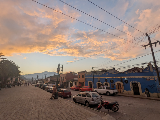 Clouds lit from below by the sunset, bright orange and pink, seen over a plaza in Mexico with mountains in the distance.