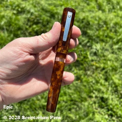 Oversized traditional styled Epic pen in beautiful Illuminated Amber Tortoise material with silver clip and clear ink window. Held in maker’s hand.