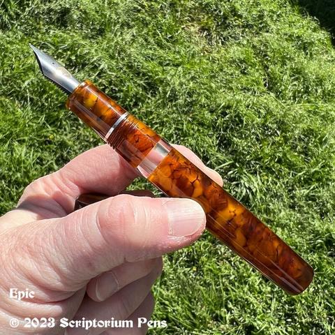 Oversized traditional styled Epic pen uncapped, in beautiful Illuminated Amber Tortoise material with silver clip and clear ink window. Held in maker’s hand.