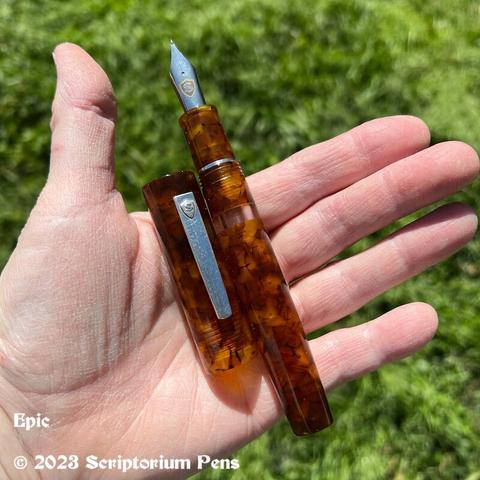 Oversized traditional styled Epic pen in beautiful Illuminated Amber Tortoise material with silver clip and clear ink window. Held in maker’s hand, uncapped, showing cap and barrel.