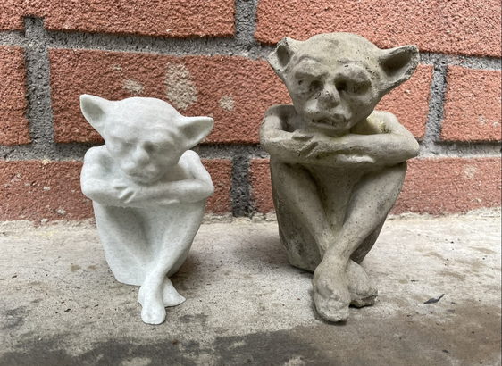 A cement gargoyle next to a roughly 3/4 heigh 3d printed version after some touch ups after the original was 3D scanned. They are sitting on cement in front of a brick wall.