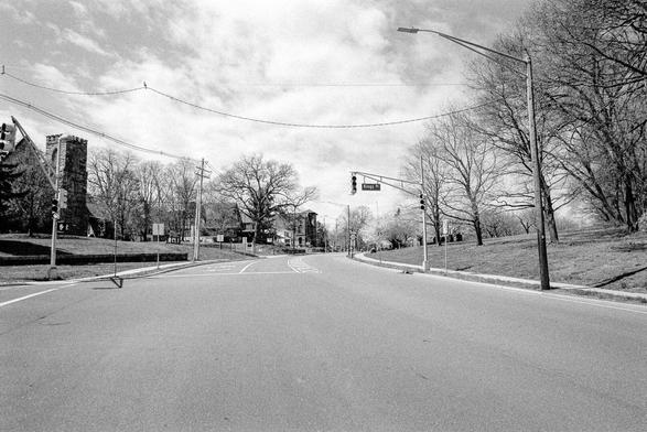 A black and white photograph showing an empty intersection with the road heading up a small hill. A traffic light hangs over the picture, along with a street sign reading “Kings Rd”. On the left, a stone church and some other houses line the street, on the right is a park with bare trees. The sky is partly cloudy. There are no signs of human activity., and the picture was taken from the middle of the road.