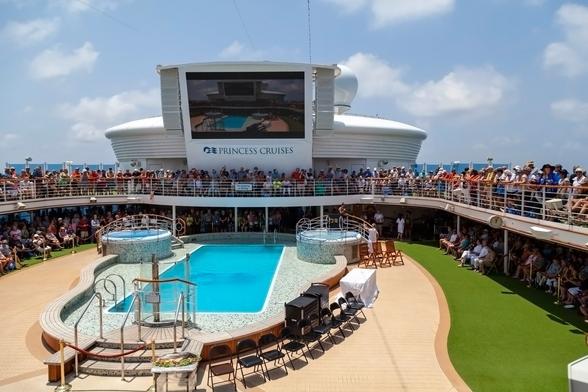 We're on a cruise ship at sea. It's a bright day with a few puffy clouds in the sky. We're by the pool deck, on the sun deck that surrounds it, and we're looking inwards towards the swimming pool and the huge outdoors TV screen that is behind and over the far end of it. All around the sun deck there are people leaning shoulder-to-shoulder on the railings and looking down. Around the pool, set back from the water and marked off with guide ropes, more people are seated and looking in. Empty chairs are set on the near side of the pool facing out, and towards one of two hot tubs at the far end of the pool there are a few more empty chairs. We're clearly getting ready to witness a popular event.