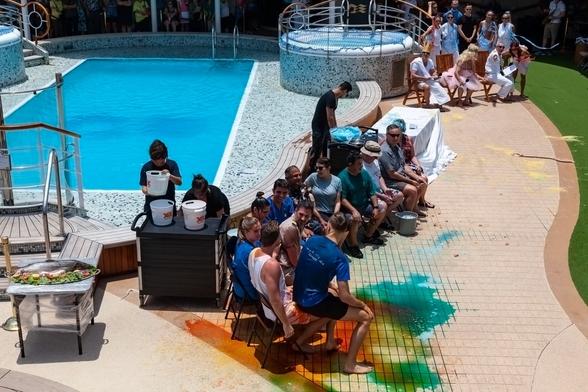 We're on a cruise ship at sea. We're by the pool deck, on the sun deck that surrounds it, and we're looking inwards towards the swimming pool, where we can see a crowd of people are gathered to watch what's happening nearer to us. Chairs are lining this side of the pool, each populated by a crew member or passenger, all of whom look wet. There's a table behind them and beyond that, beside a hot tub there are some more chairs with three people seated: the captain of the ship, barefoot, a bearded man in a blonde wig and pink dress, and another man with a long toga and a tall crown. On the ground in front of the wet people nearer us is lots of liquid in various bright colours: greens, yellows, reds, all mixing together and running into the gaps in the tiles. The people have clearly been treated to a dunking of some sort. Left of the view we can see a large fish on a table, and a couple of crew members lifting up buckets in preparation for more pouring over the assembled people. It looks a fun, party-like atmosphere.