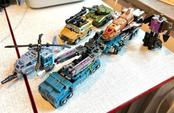 Iron Factory War Giant team in vehicle mode, a,Otley collection of military cars, trucks, tanks, a helicopter and a space shuttle. The shuttle, Blast Off, is being towed by the leader truck Onslaught, while the old G1 Blast Off stands nearby in bot mode.