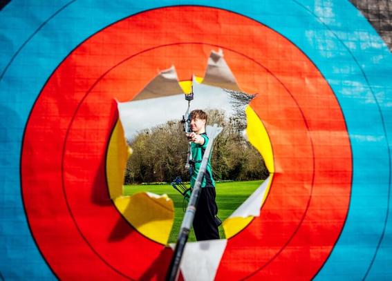 A photograph taken looking through an archery target that has been burst open. Behind the blue, red and yellow target we can see an archer in a green top and black trousers aiming at us though the hole. They are stood on a vast green, tree-lined lawn. The target goes from blue concentric circles on the outside, to red for smaller circles, and yellow for the smallest