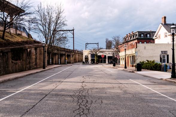 A photograph  of an empty street in a downtown commercial area in the middle of the day. Dark, closed, one to three story buildings are visible, empty parking spaces line the street, and there is no human activity.

The photograph was taken from the middle of the street towards an intersection.
Elevated train tracks run along the left side of the photograph parallel to the direction of the photograph , sitting on a grey concrete trestle. A small portion of the station platform is visible in the top left, and catenary structures holding  wires straddle the tracks at regular intervals.