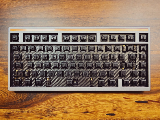 Gray 75% keyboard with copper accent bar at the top with black switches on a carbon fibre plate