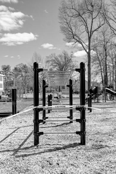 A black and white photograph of a closed school playground during COVID lockdown. The photograph shows “monkey bars” from one of the end ladders, the horizontal top bards leading directly away from the point of view. A larger set of playground equipment is in the background to the right, and the white, two-story school building is just visible to the left. Bare trees are scattered around the area.