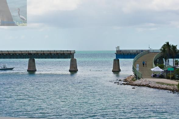 A photograph of a bridge over the ocean. The bridge emerges on the left, has a missing span, in the middle, and an intact bridge span with a wooden boardwalk down to a sandy island.