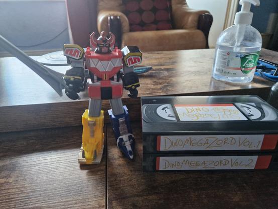Dino Megazord figure next to display stand, designed to look like two stacked old VHS tapes.