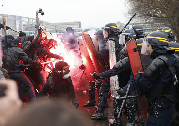 Vermummte mit Pyrotechnik in Schwarz zum Teil mit Stöcken, gegenüber behelmte Polizisten mit Schutzschildern und Schlagstock. Bild ist in Bewegung entstanden. Von oben richtet ein Mensch eine Kamera auf das Geschehen