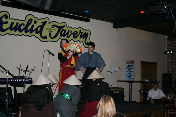 Six actors on a tavern stage perform a Japanese inspired play. Two actors stand, one hiding behind a fan, while the other four pretend to be giant mushrooms.