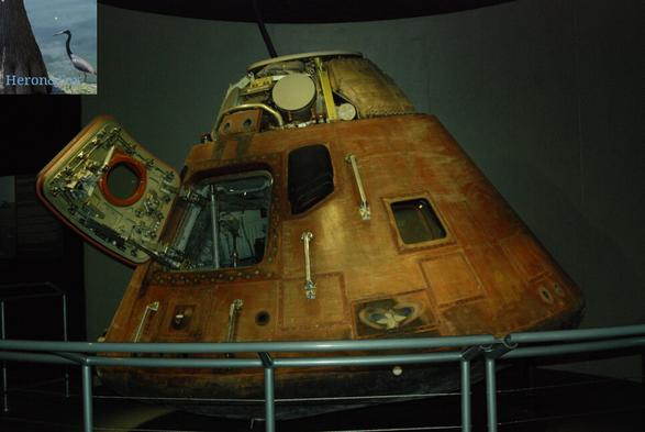 A photograph of a scorched Apollo Command Module, a cone-shape with a rounded bottom. The surface is mostly scorched brown and orange. The hatch is open, allowing the view of the complex opening system and inside the capsule. A rail occupies the foreground at the bottom of the image.A photograph of a scorched Apollo Command Module, a cone-shape with a rounded bottom. The surface is mostly scorched brown and orange. The hatch is open, allowing the view of the complex opening system and inside the capsule. A rail occupies the foreground at the bottom of the image.