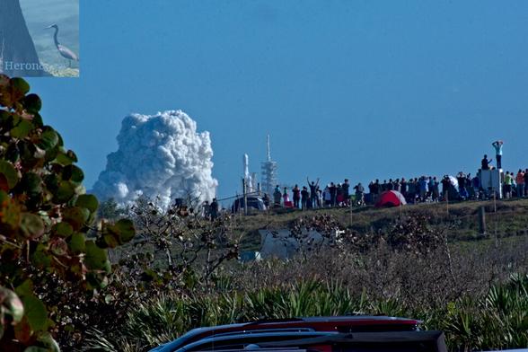 A photograph of a SpaceX Falcon 9 Heavy rocket launch. The rocket, with three first stage cores strapped together side-by-side with the upper stage and payload on the center core occupies the boundary between the top and middle vertical thirds is rising over a hill. Three jets of flame are visible from the rocket’s engine exhaust. There is a smoke cloud on the left, nearly round.  In the middle third, the top of the hill is covered in people who are watching and cheering. In the bottom third and foreground, there is the grass and scrub that covers the hill. There is a sea grape bush on the bottom left.