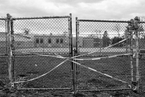 A black and white photograph of caution tape around a closed, somewhat broken gate in a chain link fence blocking access to a school athletic field. The school building is visible in the background at the far end of the field.