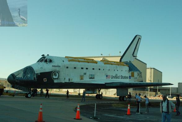 A photograph of NASA’s Space Shuttle Atlantis, occupying the vertical middle third of the image. The Shuttle is missing part of its nose, which is covered in clear tarp. The two, white orbital maneuvering system pods are missing. It is being towed along the ground. Two brown hangers are in the background.