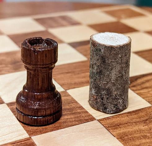 a black rook on a chess board, with an uncarved wood stick beside it.
