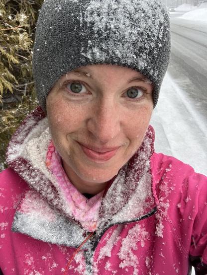 Selfie of a middle aged white woman post run. There's snow on her toque, her buff, her hood, and her bright pink jacket. The background is snow covered. She has an amused smile on her face.
