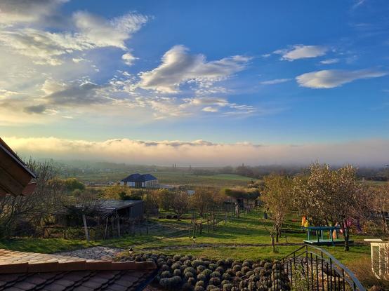 Photo of a landscape in early morning. The sky is blue, dotted with clouds, and there is a line of clouds just above the horizon as if they were mountains in the distance. In the foreground there is a garden with almond trees in bloom, green grass, lavender bushes, and a stone garden path. Colorful fabrics hang from the trees as decoration (for an activity this morning). There are vineyards in the distance.