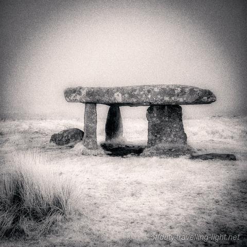 Toned black and white infrared photo of a chambered tomb with a capstone on three uprights and a misty bacground.