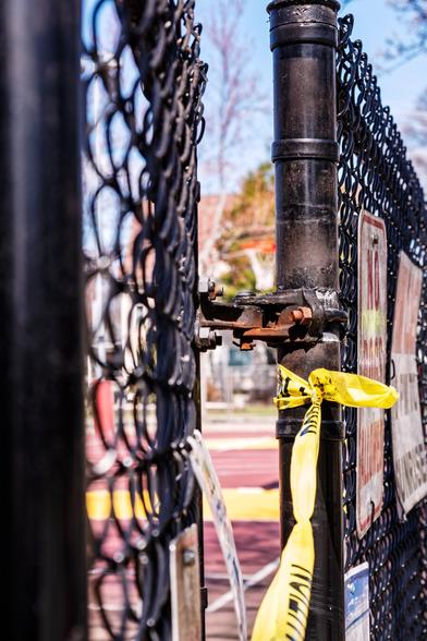 A color photograph of a closed  gate in a black chain link fence. The focus is on the hinge on the fence pole, with the gate Itself to the left and the basketball court in the background blurred. Yellow “Caution” tape is tied to the post and draped across the gate.