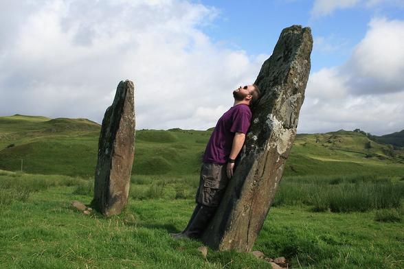 A white man with a beard leaning against a standing stone. The stone is at quite an angle. Beyond is a second stone, and in the background, hills. The sky is blue with white clouds, the sun is shining.