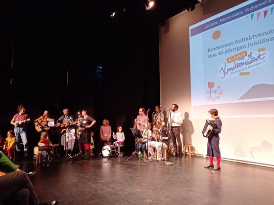Auf der rechten Seite ist ein Beamer -Screen, auf dem steht: "Kindernest-Auftaktveranstaltung zum 40-jährigen Jubiläum". In der Mitte des Bildes sieht man die Kindernest-Band: eine bunte Gruppe an Leuten, Eltern ind Kinder, mit bunter Kleidung und unterschiedlichen Instrumenten. Sie gestalten das Rahmenprogramm und unterhalten mit ihrer Musik das Publikum gut. Mitgesungen werden muss selbstverständlich auch!