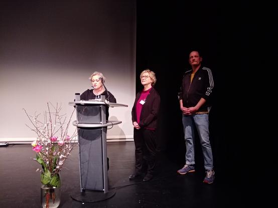 Eine Person steht am Rednerpult: es ist Sylvia Leippert-Thomas, die Gründerin und pädagogische Leitung des Kindernests. Neben ihr Rita Herzog, die den kaufmännischen Bereich betreut. Beide sind festlich gekleidet. Neben ihnen steht Manuel Deuse, ein großer Mann mit Schnauzer und Sportjacke, der Vorsitzende der Elterninitiative. Ein Blumenstrauß ist auch zu sehen. Es ist alles sehr festlich.