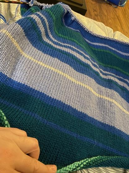 Eine gestrickte Decke. Die Decke ist gestreift in unterschiedlichen Blautönen. Die Streifen sind unterschiedlich breit. Am oberen Ende ist eine Stricknadel zu sehen.