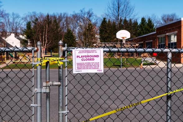 A color photograph of a closed gate in a chain link fence  taped off with yellow “Caution” tape. Beyond the pavement and basketball hoop, a playground and grass, and a portion of a school building are visible.

Attached to the gate is a sign that reads “AS PER MORRIS COUNTY EXECUTIVE ORDER NO. 104, PLAYGROUND CLOSED. PLEASE BE ADVISED THST AS OF WEDNESDAY MARCH 18, ACCESS TO THIS PLAYGROUND SHALL BE RESTRICTED AND SHALL NOT BE PERMITTED FOR ANY PURPOSE.”