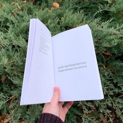 The book Capsule Stories Winter 2022 Edition held open to the poem “[swan song]” by Katelyn Grimes in front of evergreen bushes