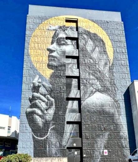 Streetartwall. On the outer wall of a six-story building is sprayed / painted a huge portrait of a woman in gray and dark gray. 20 meters high and 8 meters wide, a magnificent mural with a sad background.
She is depicted sideways, has long hair and her eyes closed. Her hands seem to be praying at first glance, but they are strangely twisted fingers inside each other. A bright yellow sun wreath appears around her head.
JDL: "It is a tribute to a friend who suffers from neuromuscular disease. I want to show that what she is is inside her, it doesn't need hands." (Le Télégramme). A tribute that conveys all the more emotion because it is on one of the walls of the emergency room of the hospital of Morlaix.
