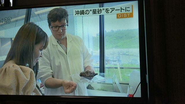 Very serious scientist (wearing a lab coat) presenting samples to TV host.